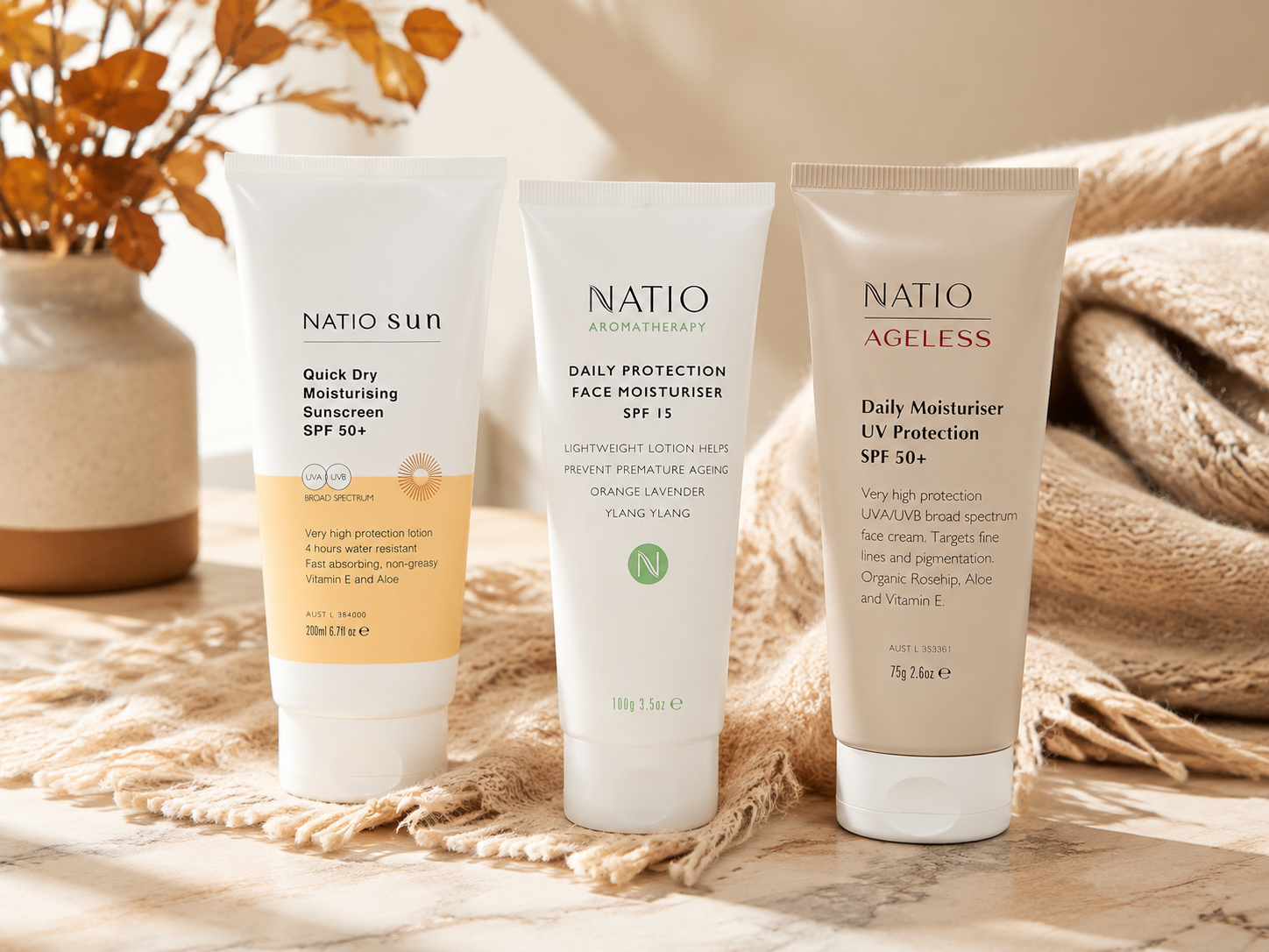 Three Natio skincare products on a light marble surface with a neutral textured cloth and dried leaves in the background, featuring Quick Dry SPF 50+, Daily Protection SPF 15, and Ageless Daily Moisturiser SPF 50+